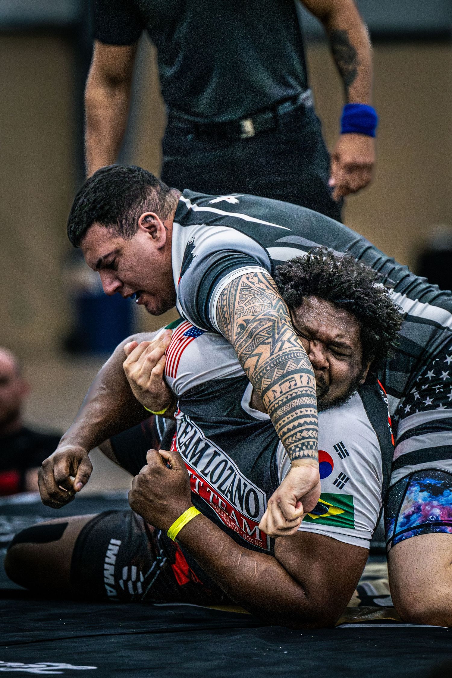 Two male grapplers compete in an intense submission wrestling match at an indoor martial arts tournament.
