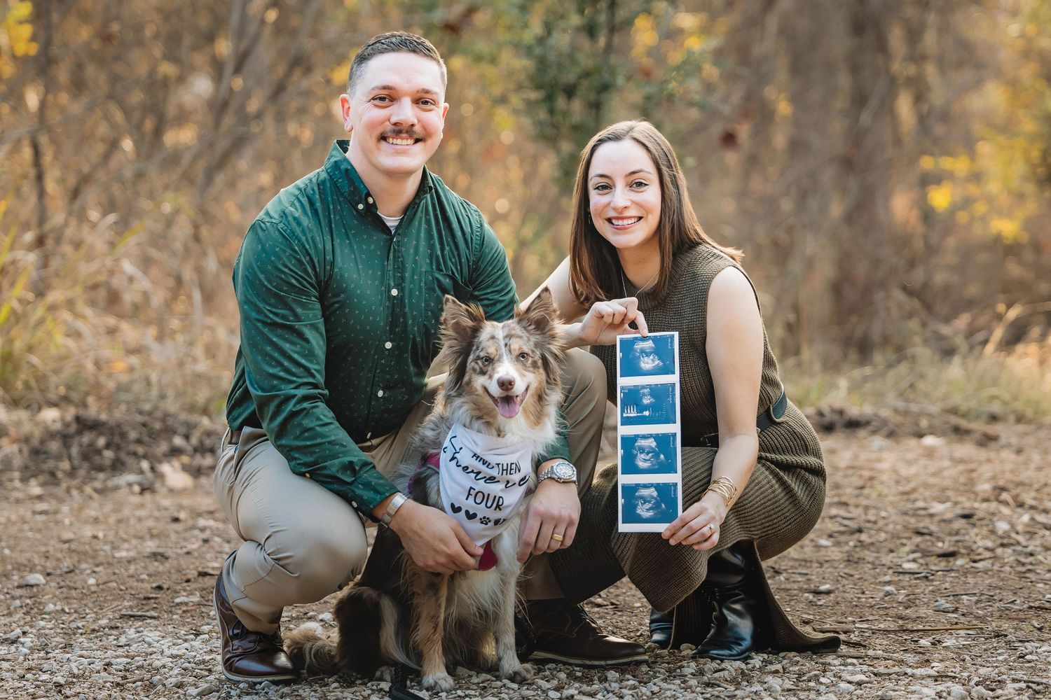 A couple poses outdoors with their small dog while holding a blue and white sign during an autumn photoshoot.