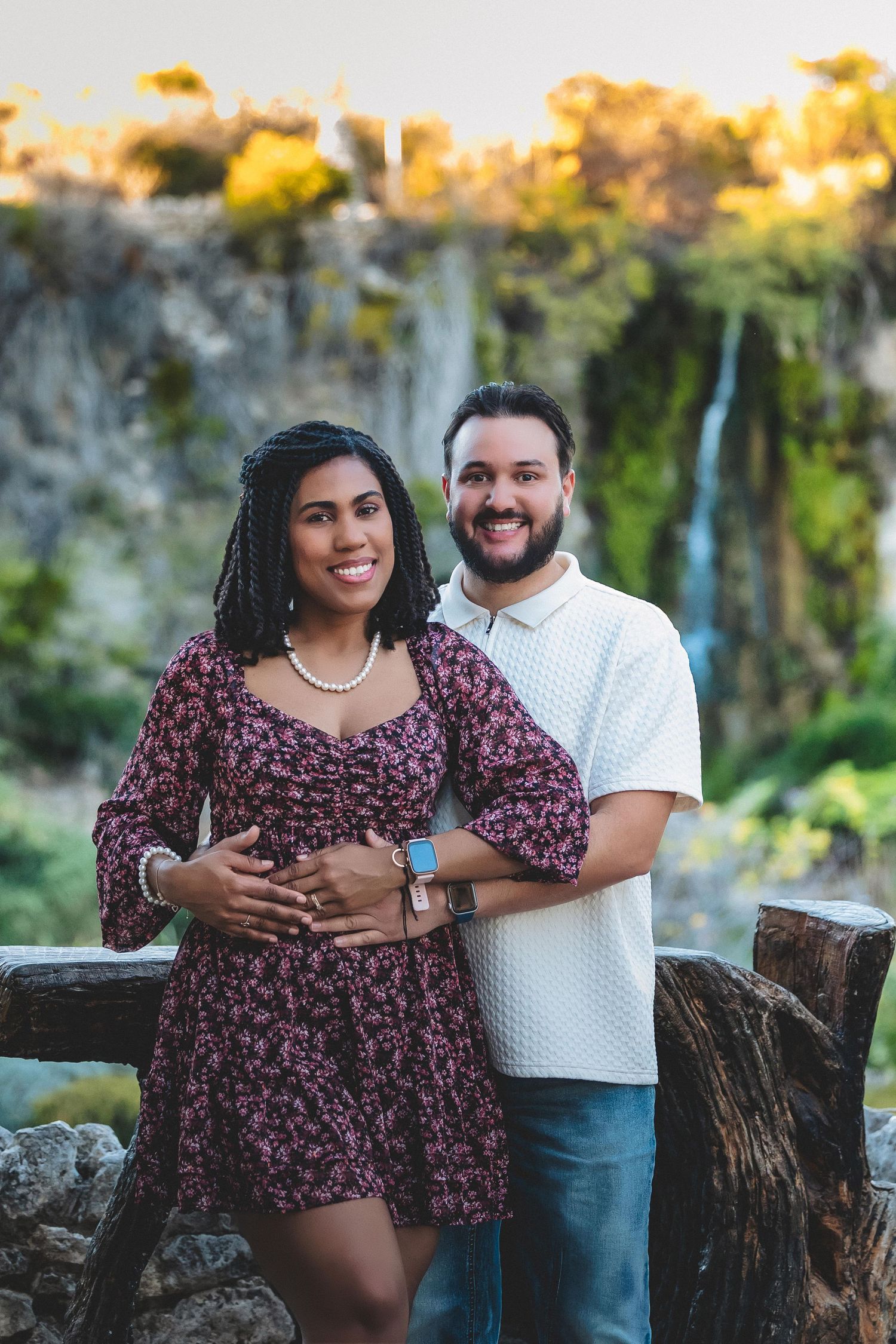 A couple poses together outdoors at sunset against a natural backdrop of trees and greenery.