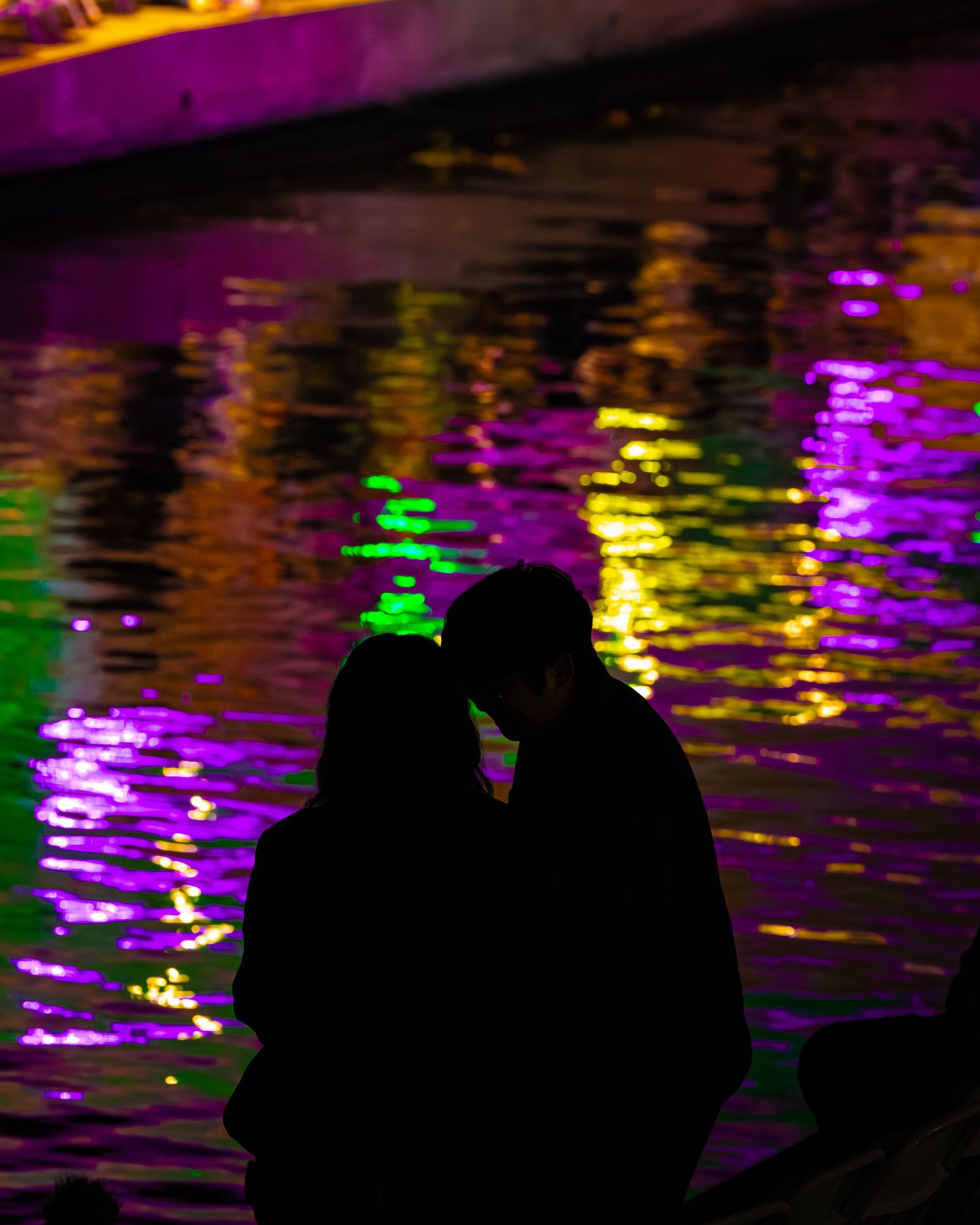 Silhouettes against colorful purple, yellow and green reflections on water at night.