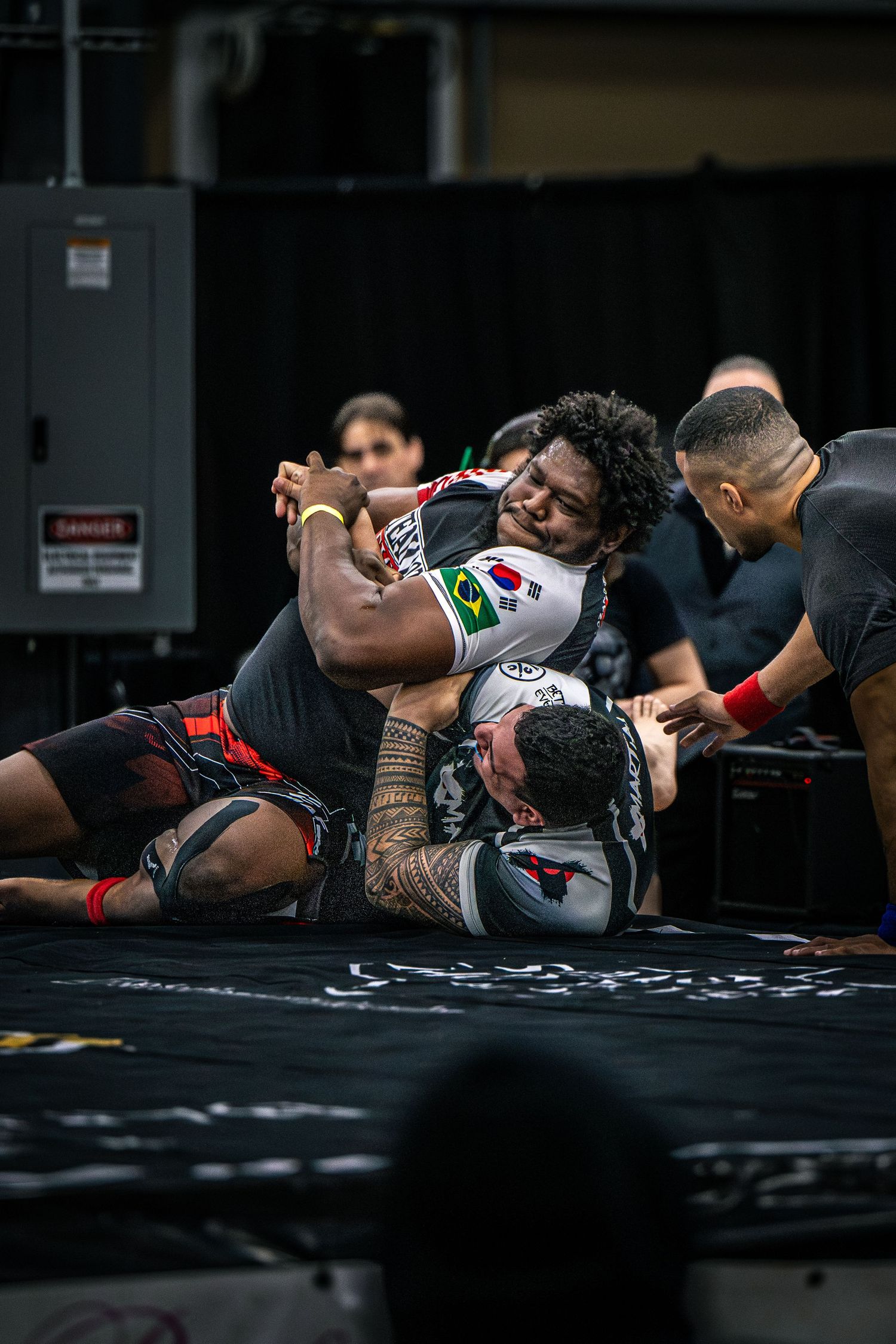 Two MMA fighters grappling on a black mat during a competitive submission wrestling match.