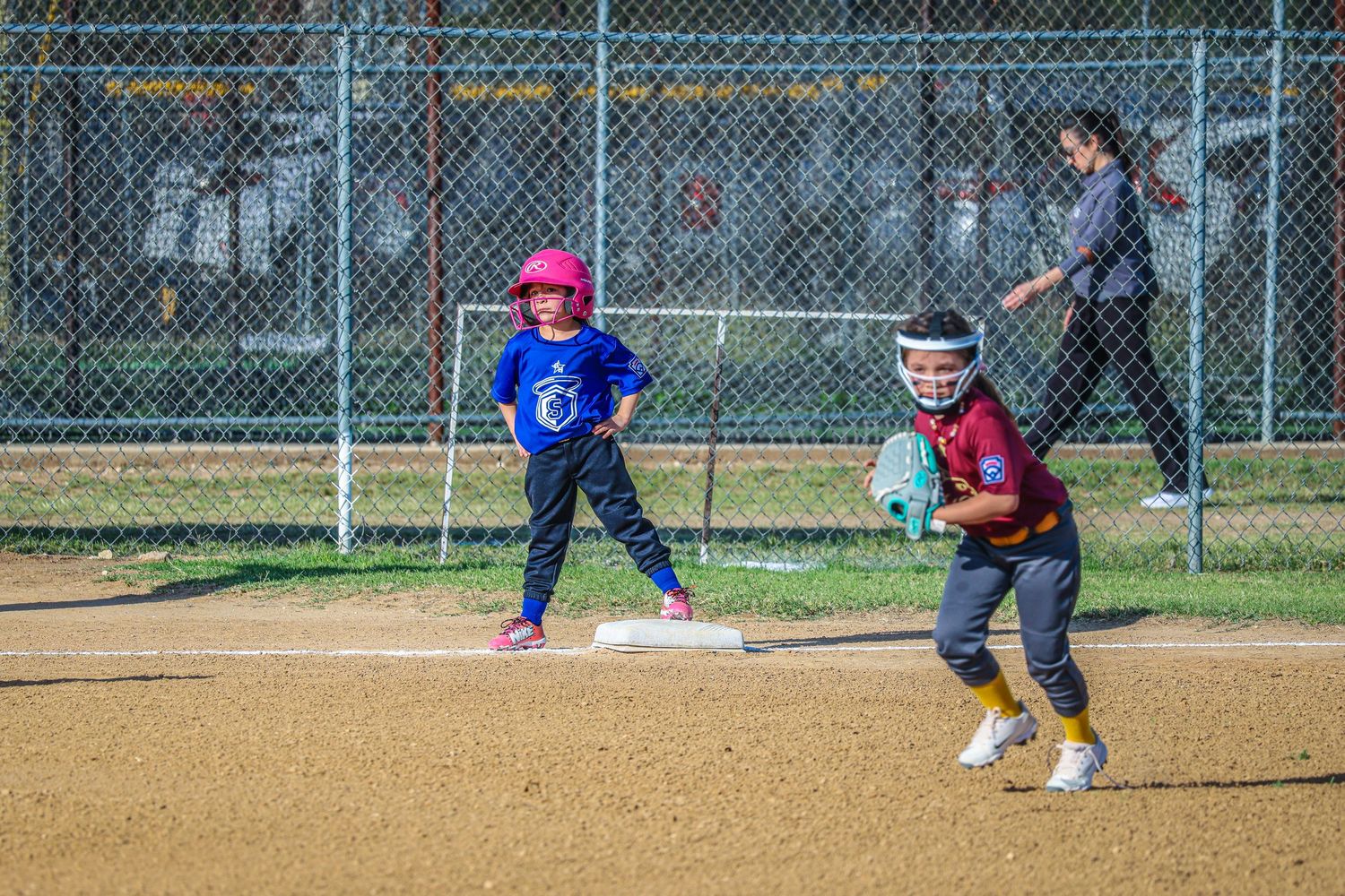 Two young girls playing softball on a dirt field, one standing on base in blue jersey, another fielding in maroon uniform.