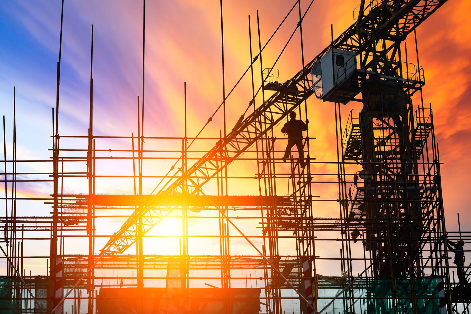 Silhouette of construction workers on scaffolding at sunset with vibrant orange and blue sky backdrop.