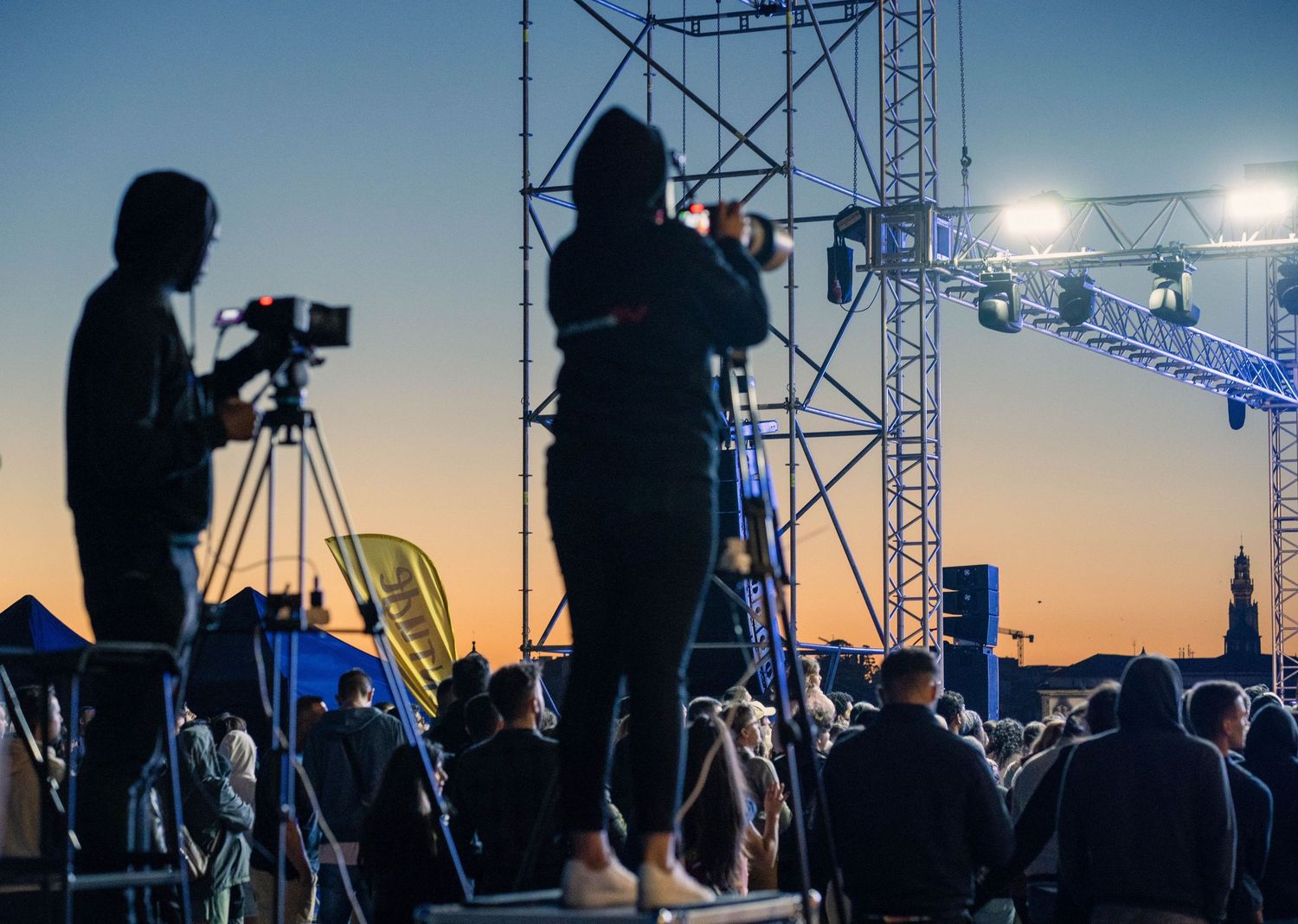 Silhouetted camera operators filming an outdoor evening concert event with stage lights and crowd in background.