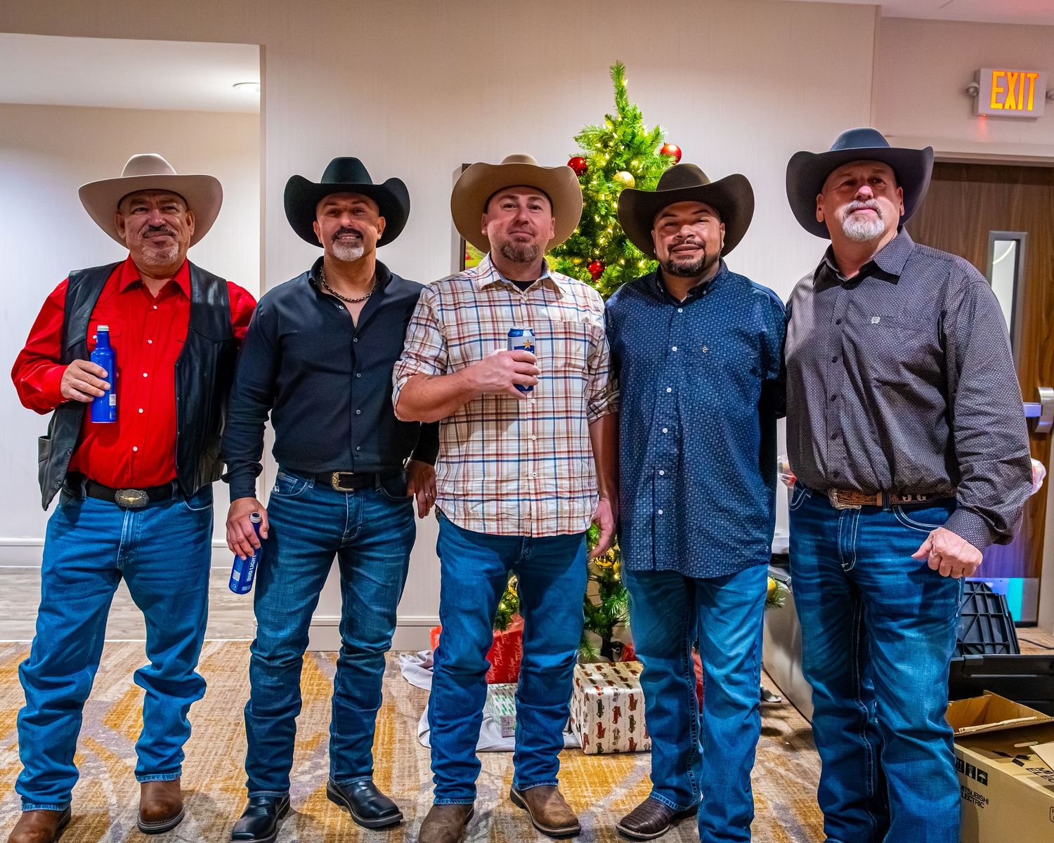 Five men in cowboy hats pose together at a holiday party near a Christmas tree.