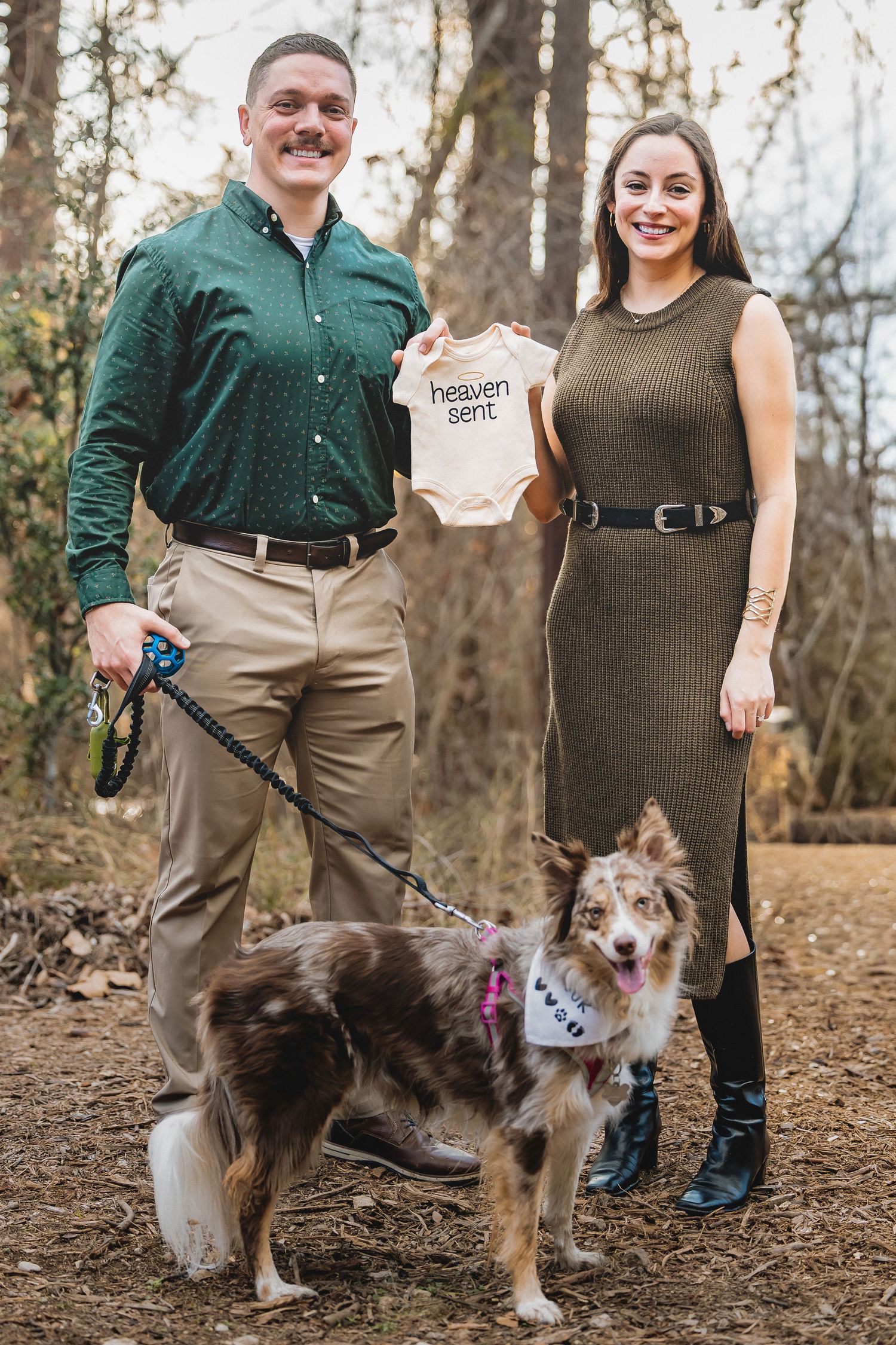 A couple poses outdoors with their Australian Shepherd dog while holding a small baby onesie in an autumn setting.