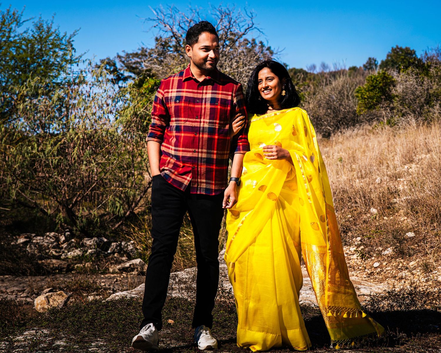 A couple in a red plaid shirt and bright yellow dress pose together outdoors against a natural autumn backdrop.