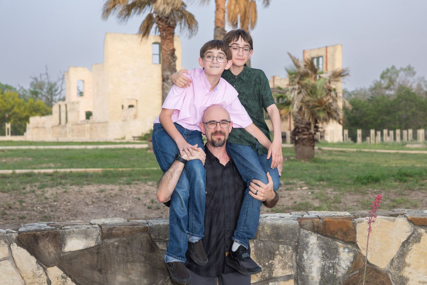 A man kneeling outdoors with two boys on his shoulders, posing near palm trees and a historic stone building.