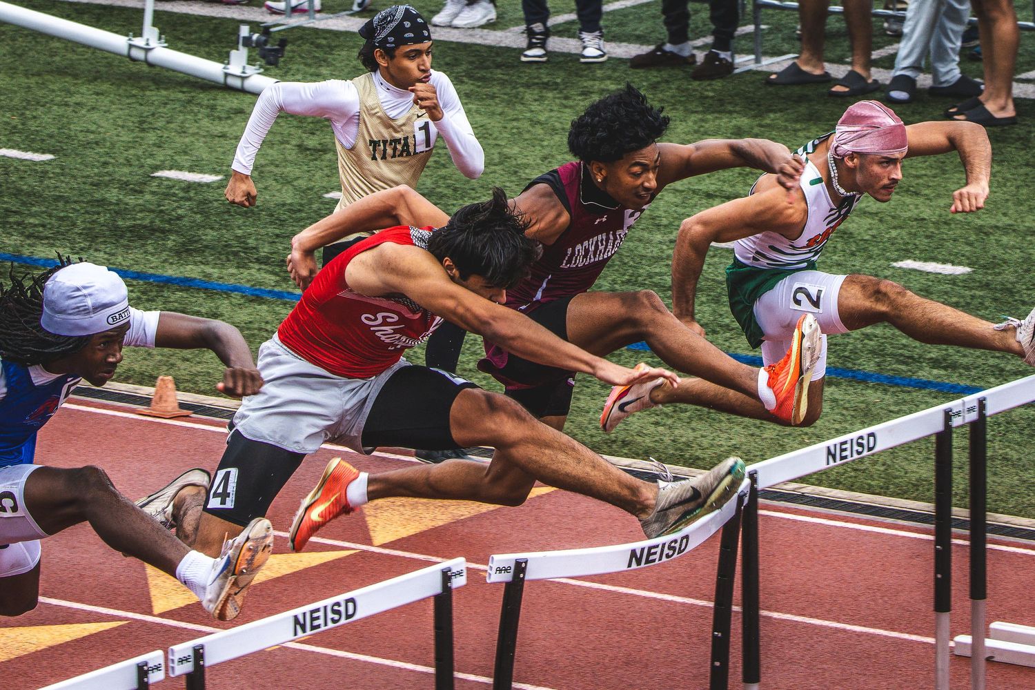 Athletes compete in a hurdles race on a track, leaping over NEISD barriers during an outdoor track and field event.