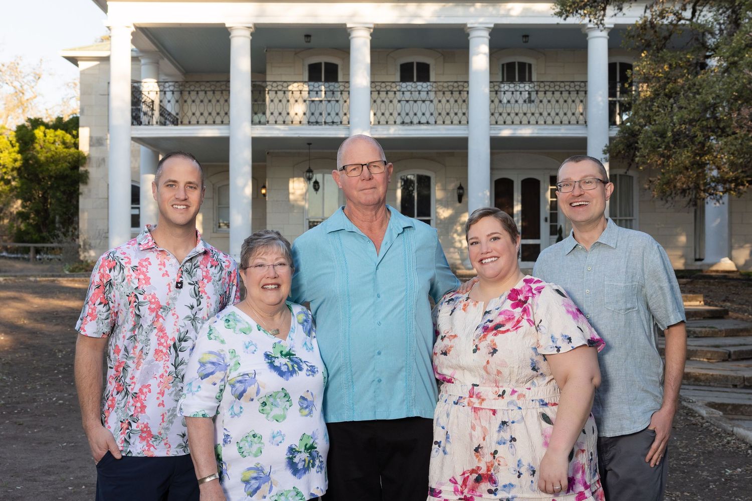 Family of five smiling together outdoors in front of a white colonial-style mansion with columns.