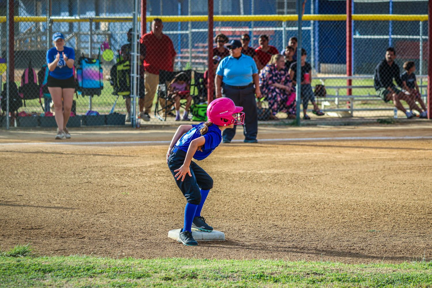 Young softball player in pink helmet crouches at base during youth league game on dirt field.