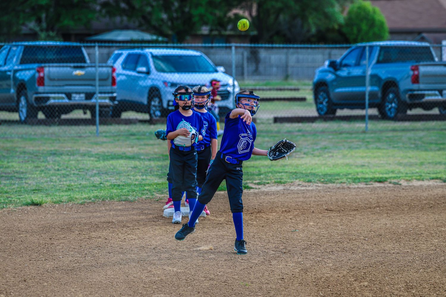 Youth softball player pitching during a evening game on a dirt infield with teammates and parked cars in background.