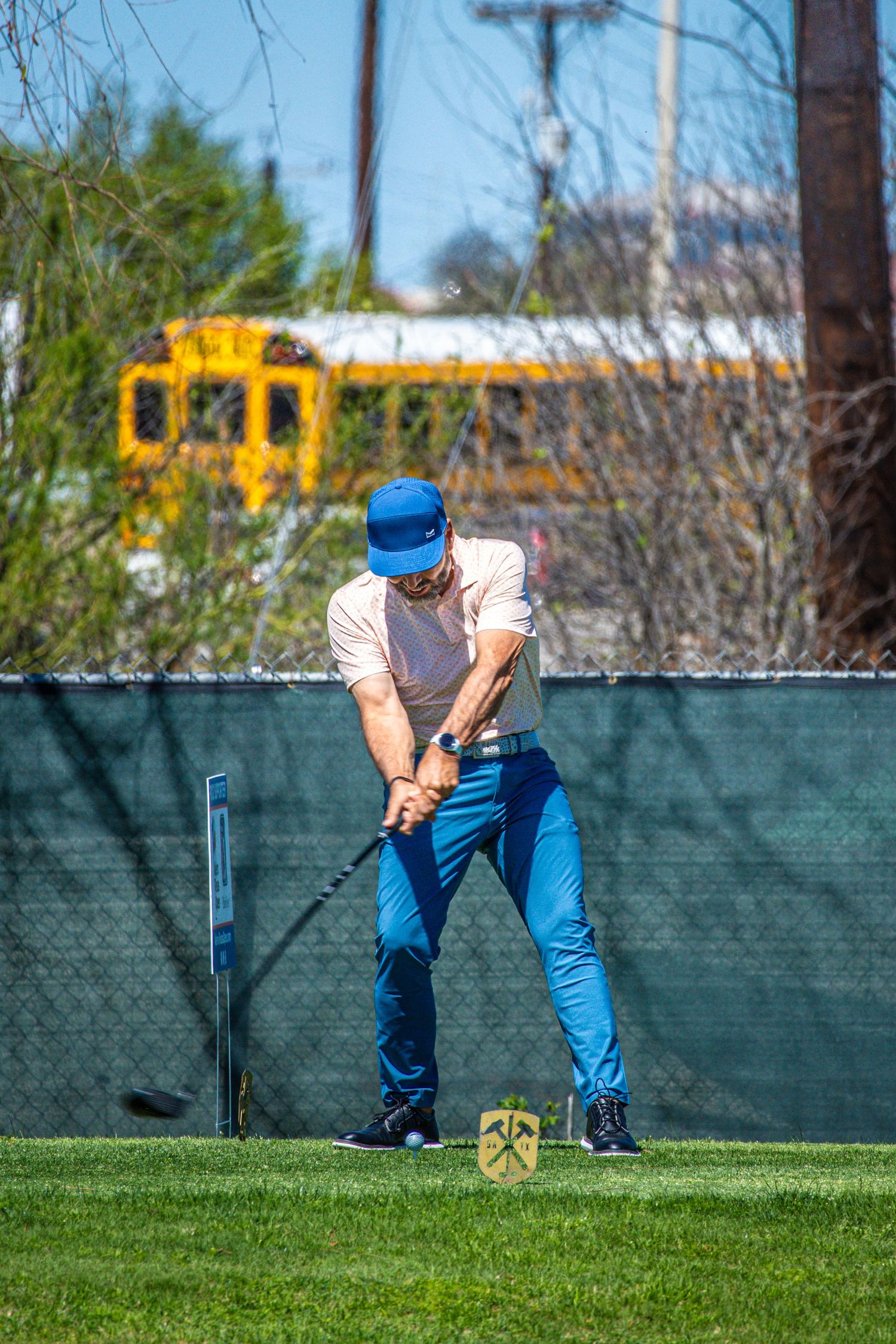 Man in blue pants and cap swinging a golf club on a green course with a yellow school bus in the background.