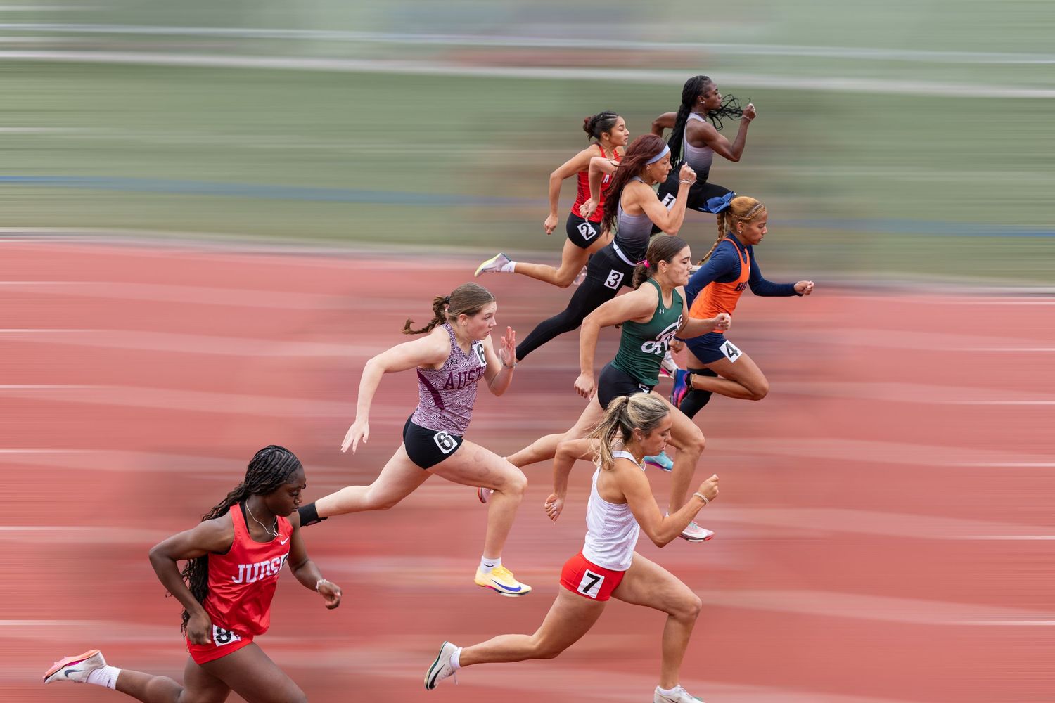 Female athletes sprint on a red track during a competitive race, captured with motion blur effect.