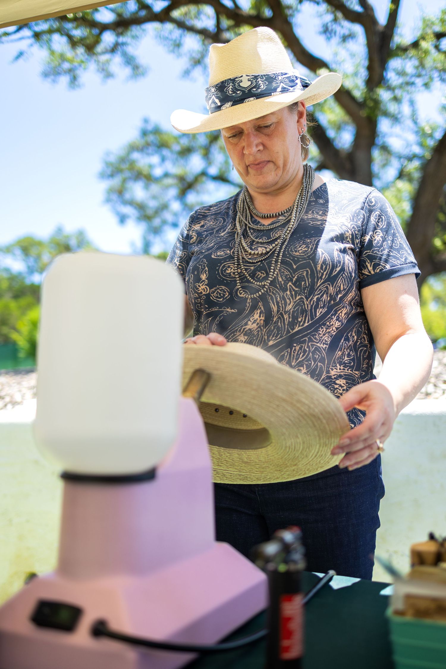 Man in cowboy hat and patterned shirt examining a straw hat at an outdoor market vendor booth.