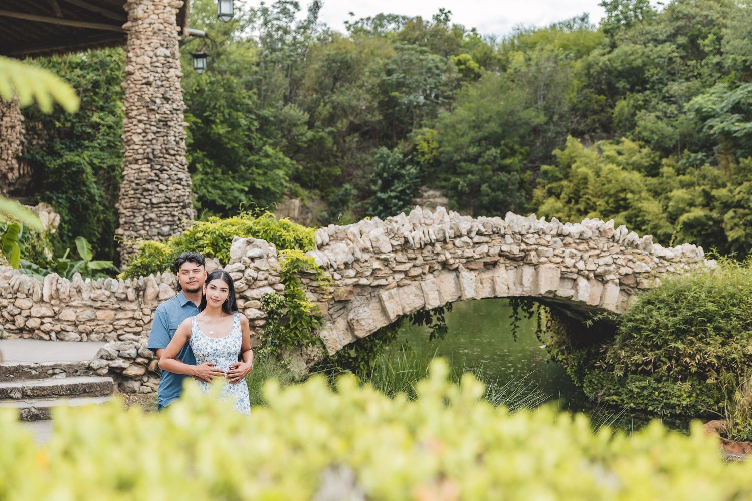 Engaged couple posing in lush garden near stone arch bridge outdoors.