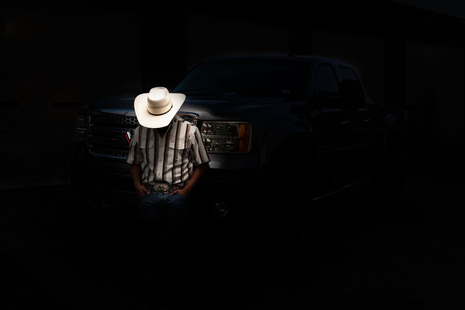 A person in a striped shirt and white cowboy hat stands illuminated in darkness before a dark truck.