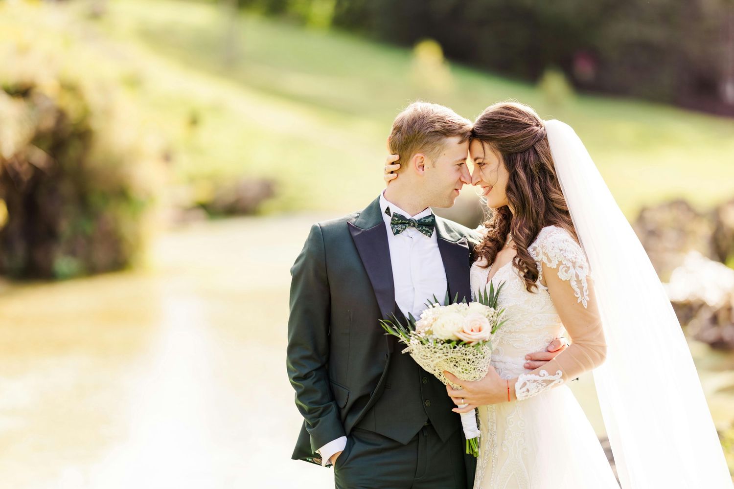 Wedding couple poses in a sunlit garden setting with white floral bouquet.