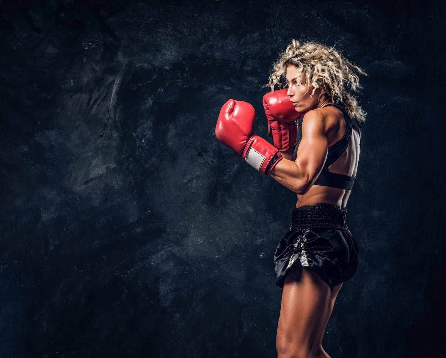 Muscular female boxer in red gloves throwing a punch in a dark studio, showcasing strength and athletic form.