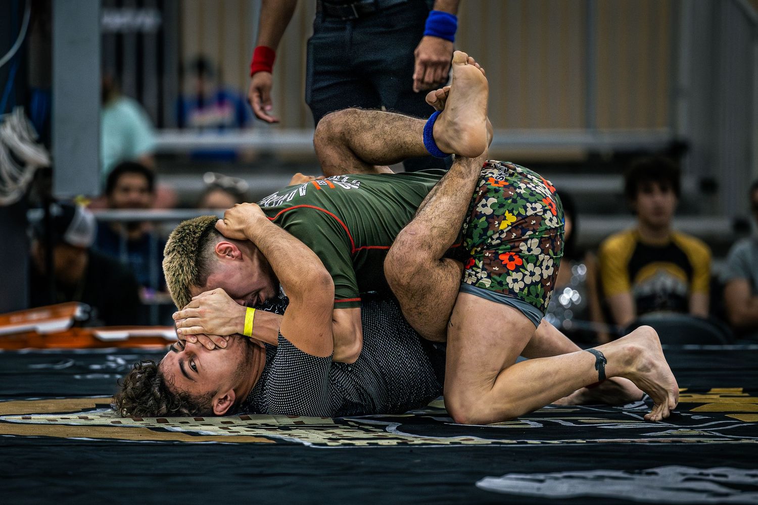 Two wrestlers grappling on a mat during a competitive wrestling match in a crowded gymnasium.