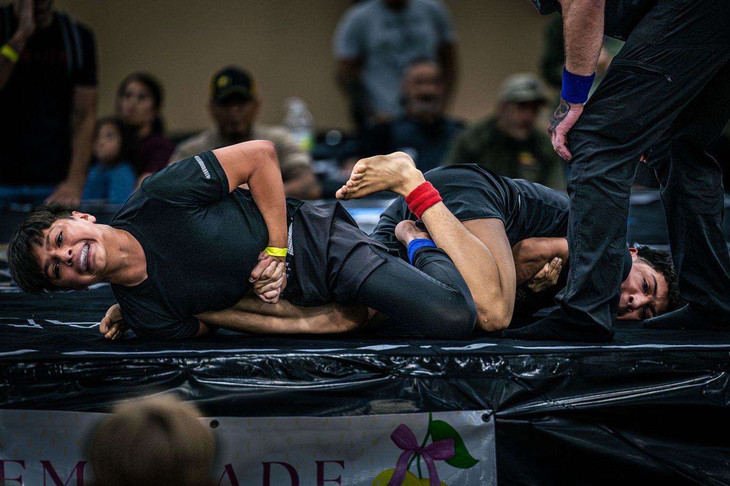 Two competitors grappling on a mat during a submission wrestling tournament in a sports venue.