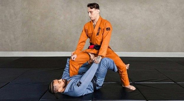 Two practitioners training BJJ, one in orange gi applying ground control over partner in blue gi on black mats.