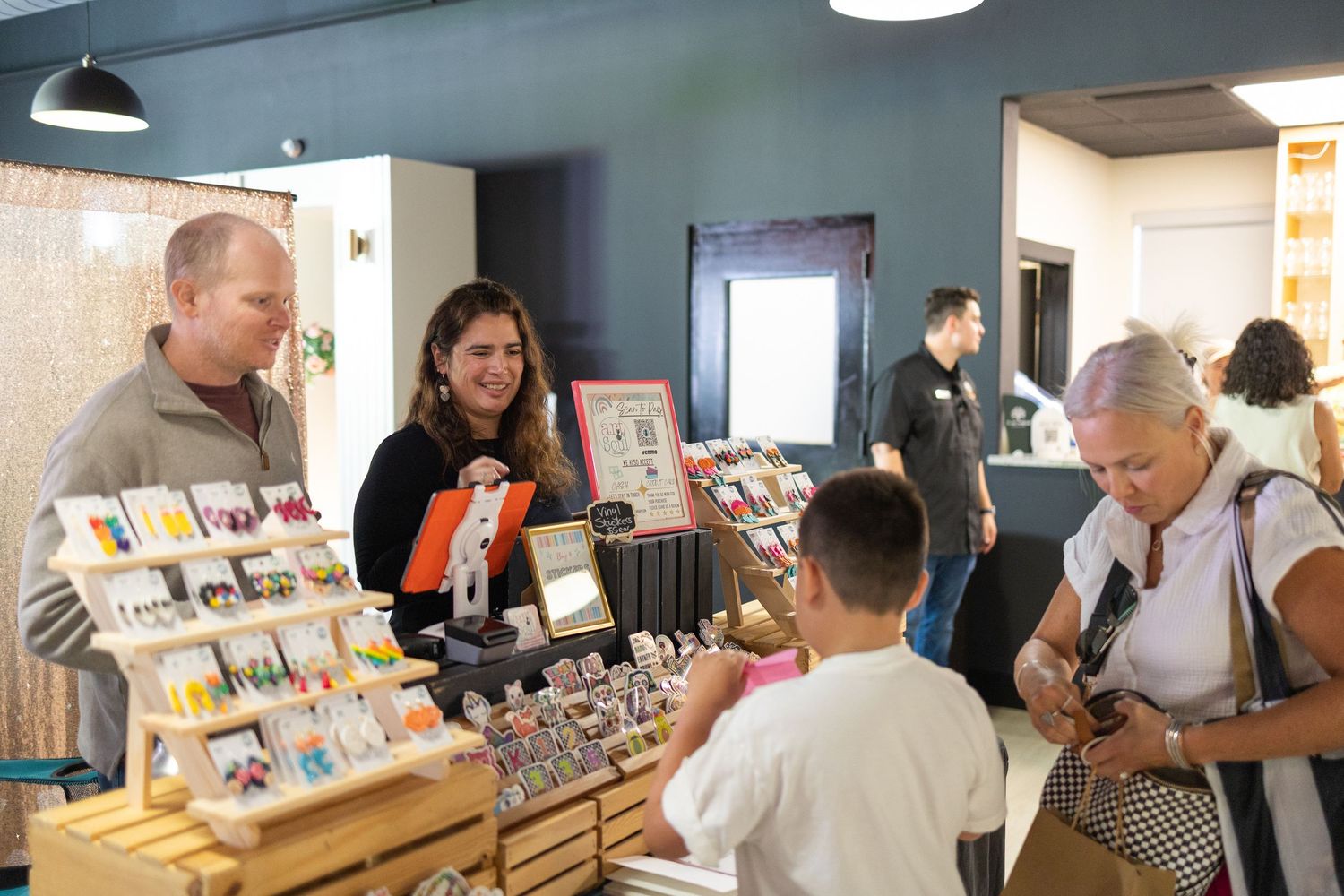 Customers browsing colorful handmade crafts and jewelry displays at an indoor artisan market vendor booth.