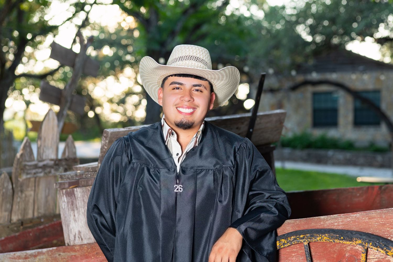 Young man in graduation gown and cowboy hat smiling outdoors near rustic wooden wagon.