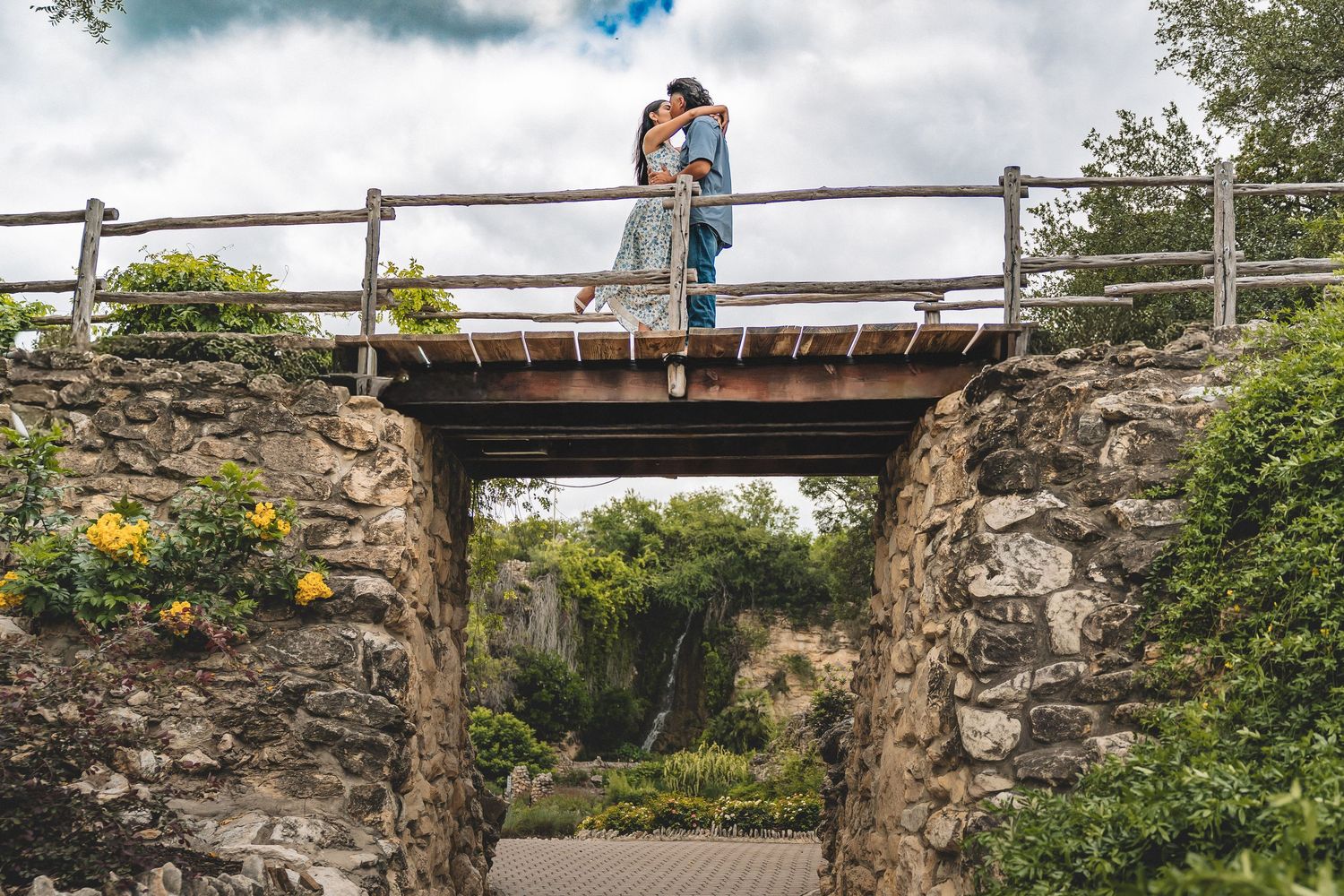 Couple sharing a romantic kiss on a rustic wooden bridge over a stone archway with a waterfall backdrop.