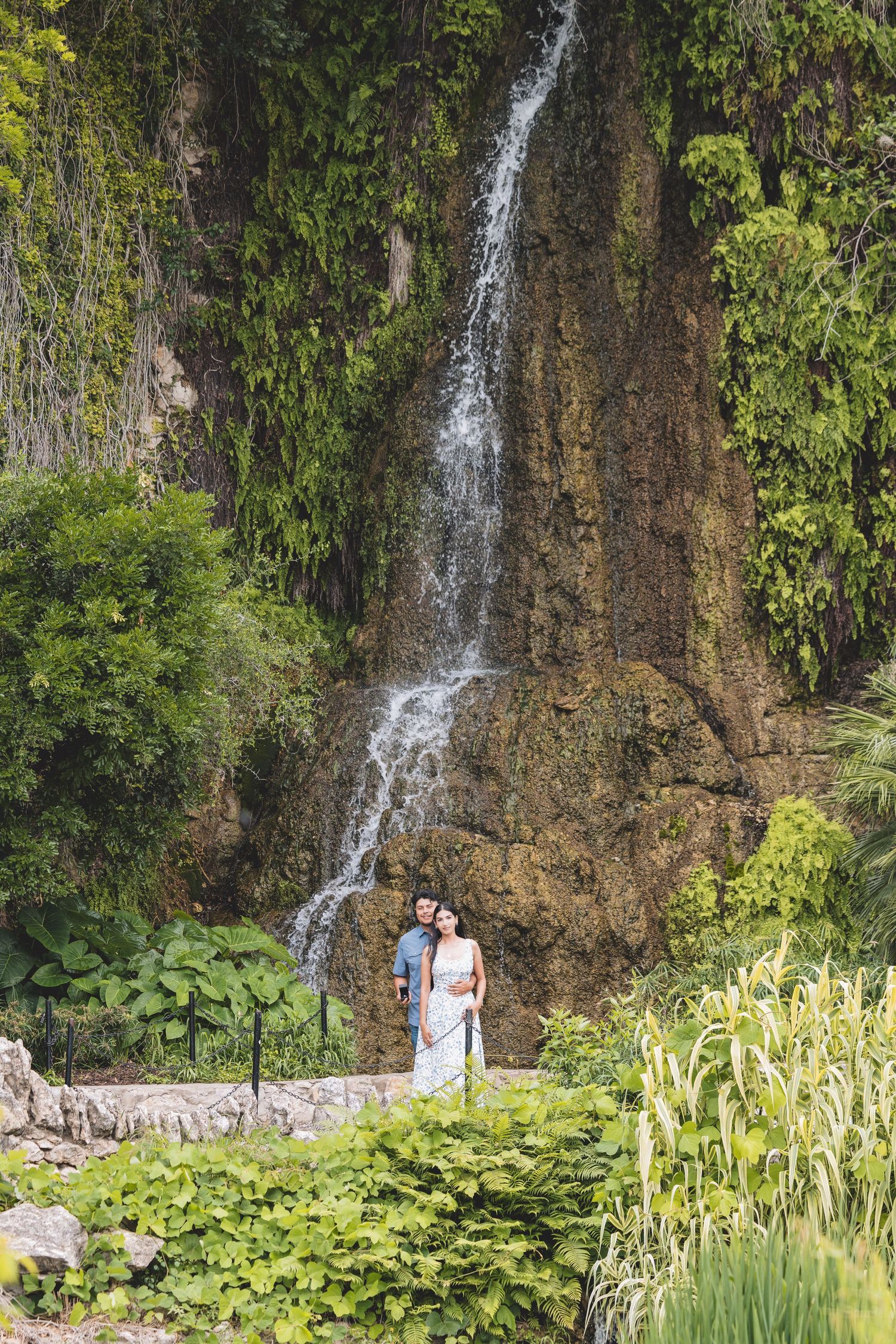 Couple embracing in front of a lush waterfall cascading down a rocky cliff surrounded by tropical greenery.