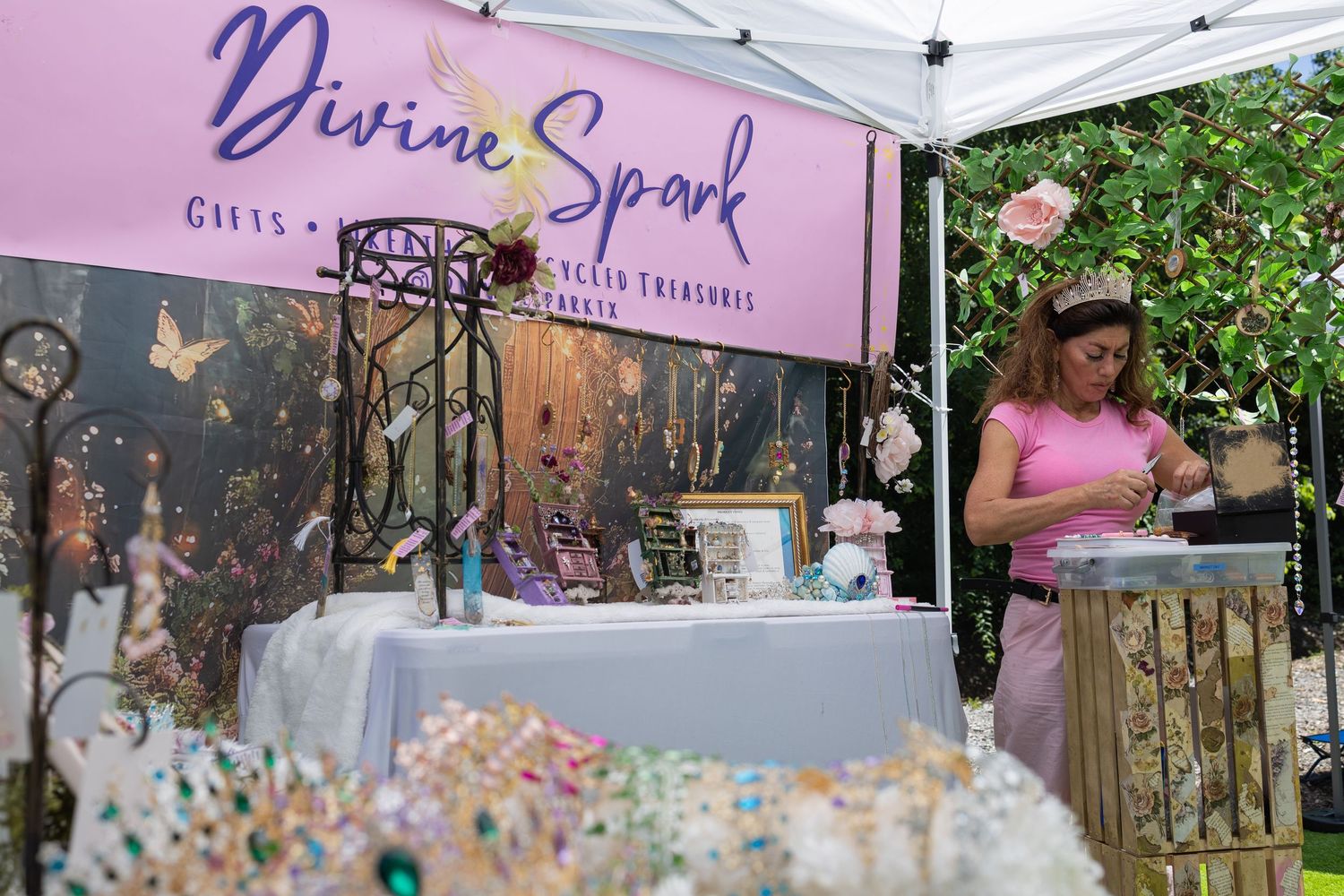 Woman arranging products at Divine Spark Gifts booth at outdoor market with colorful wands and fairy treasures on display.