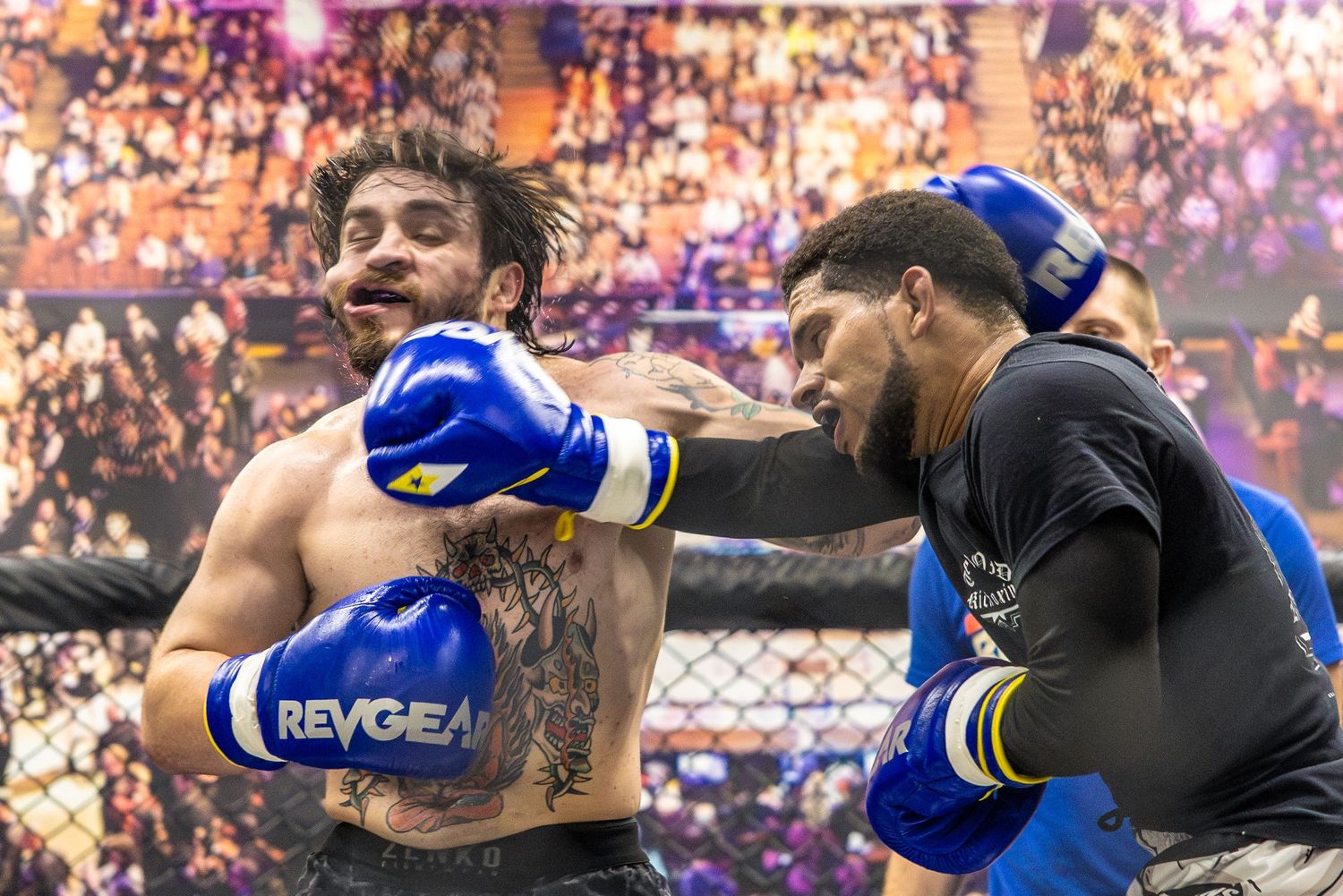 Two tattooed male fighters exchanging punches wearing Revgear blue boxing gloves in a ring.