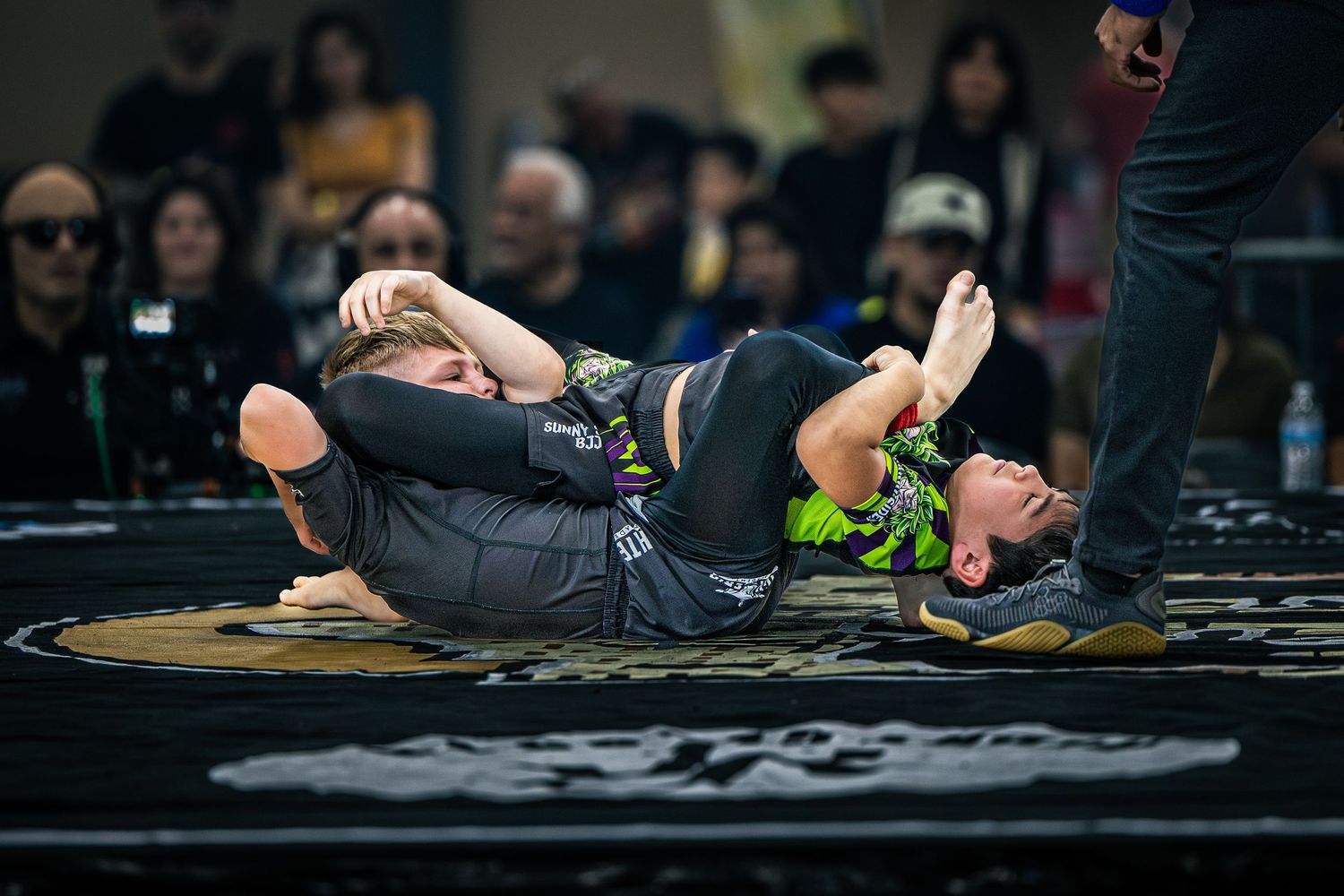 Two athletes competing in a grappling match on a black mat at an indoor martial arts tournament.