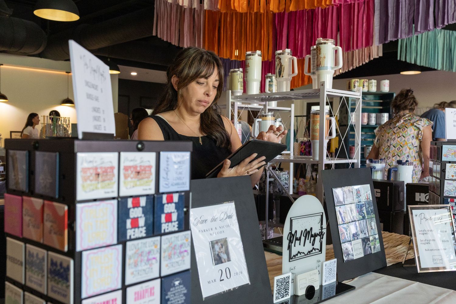 Woman using tablet at craft market vendor booth with colorful yarn display and product cards.