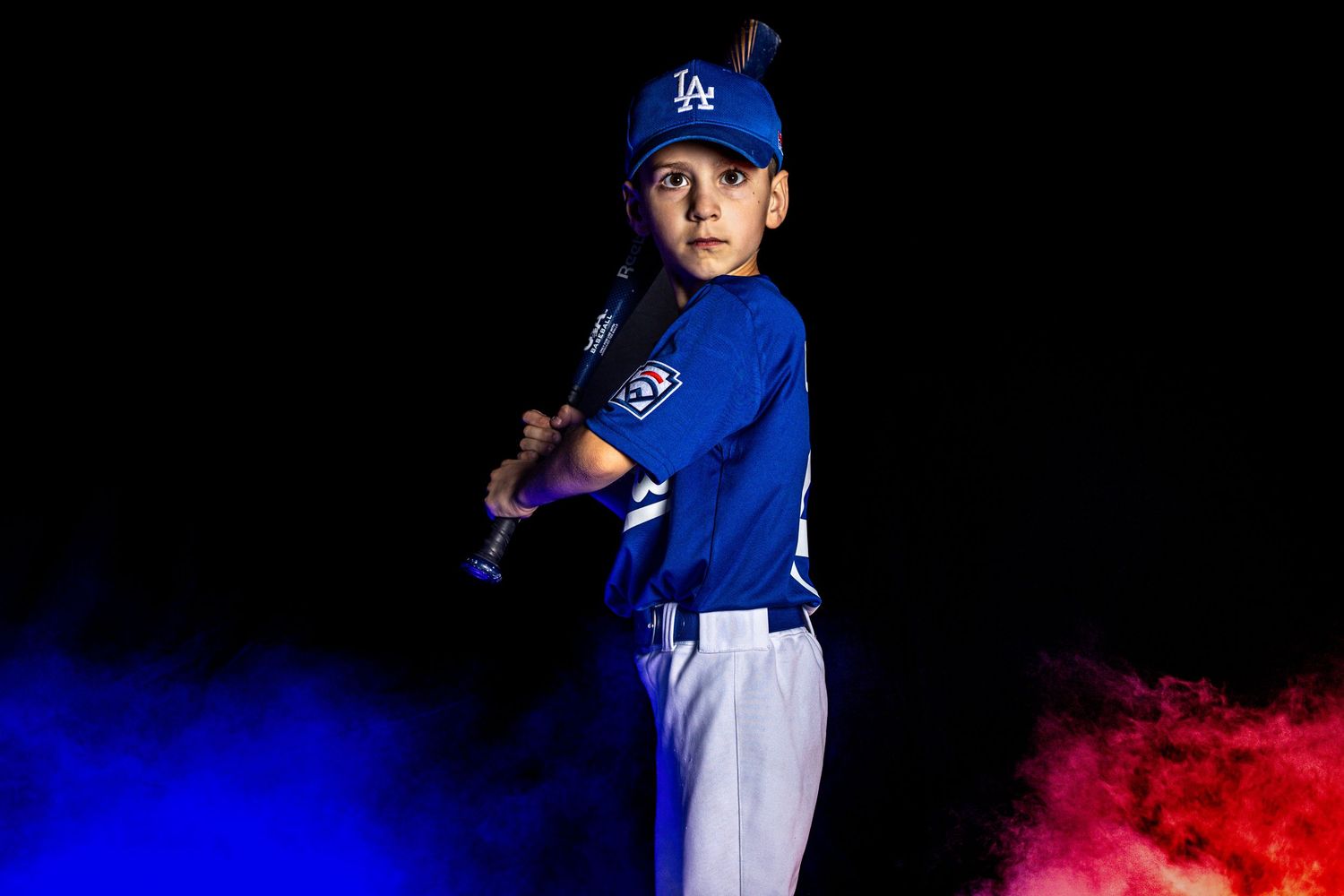 Young baseball player in LA Dodgers uniform holding bat, posed dramatically against dark background with colorful smoke.