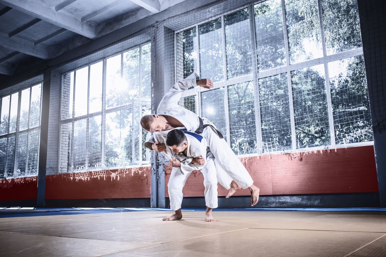 Two judo athletes in white gi perform a dynamic throwing technique during training in a gym.