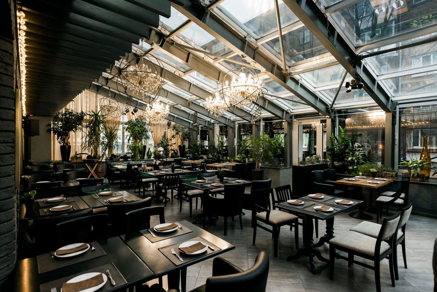Elegant restaurant with glass ceiling, dark tables, lush plants, and chandelier lighting in a modern dining space.