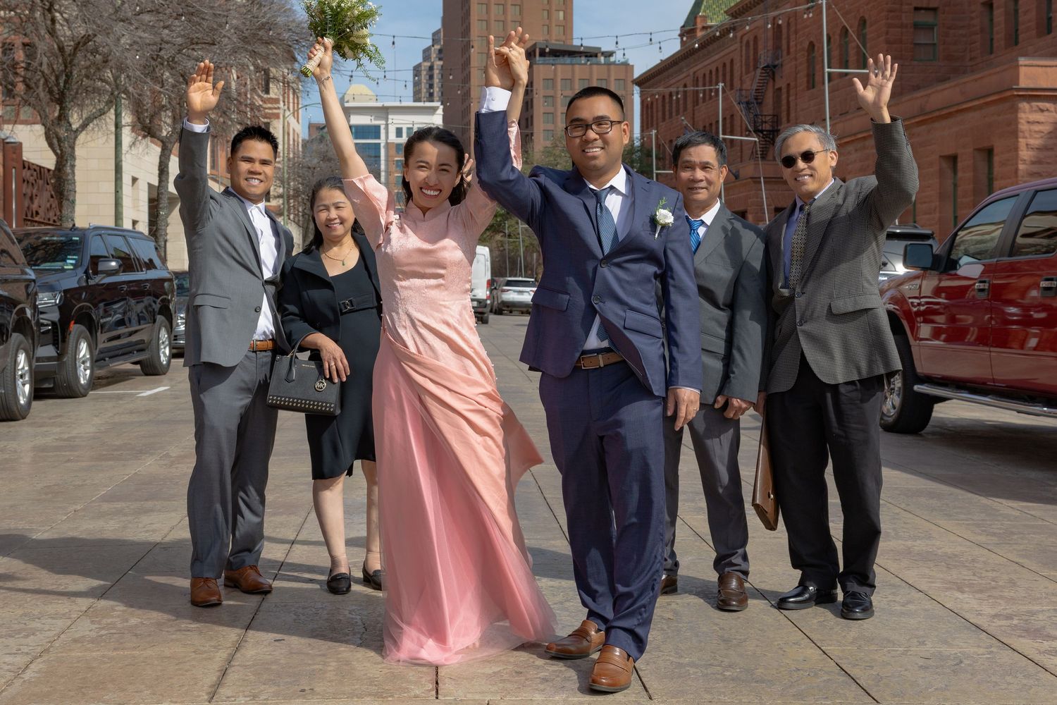 A group in formal attire poses together outside a brick building on a sunny day.