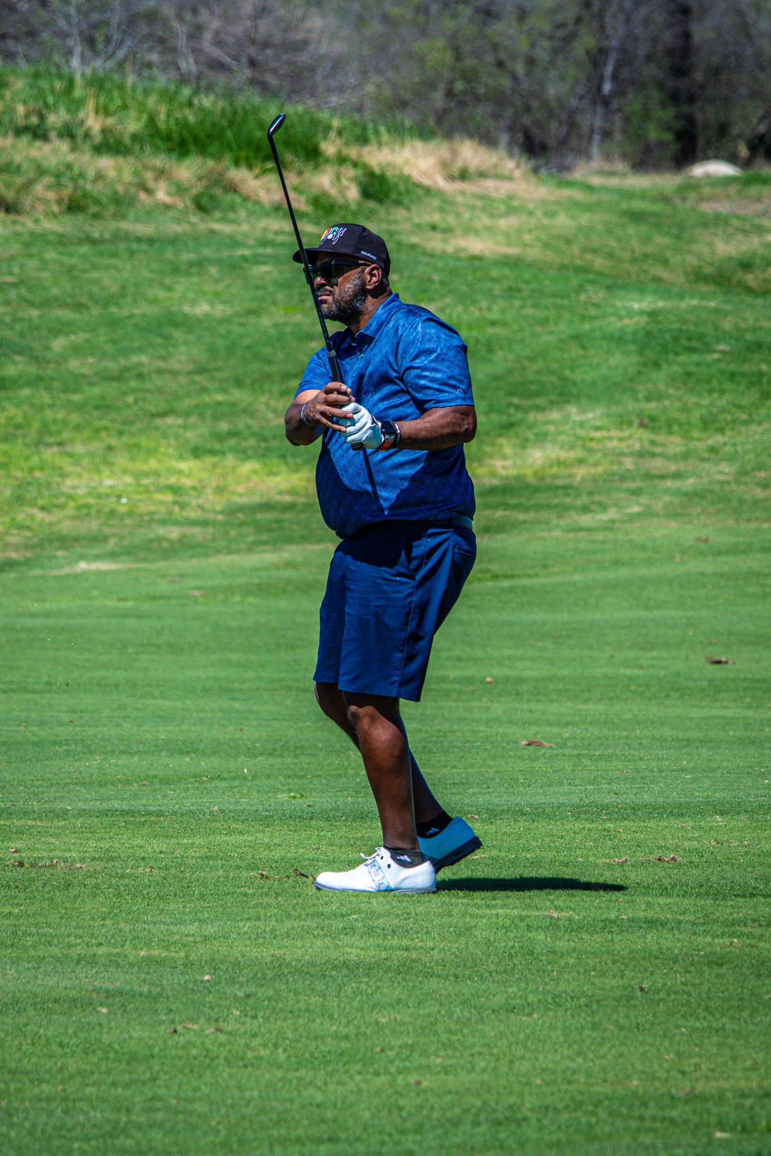 A man in blue shirt and shorts follows through on a golf swing on a lush green fairway.