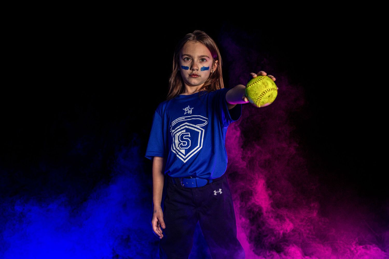 Young girl in blue softball uniform holds yellow ball amid colorful smoke on dark background.