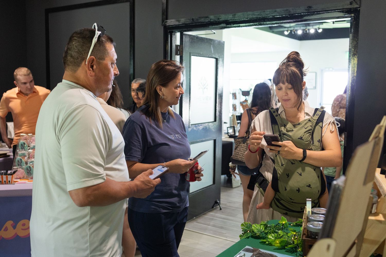 People browsing and discussing products at an indoor vendor booth with fresh greens displayed on a table.