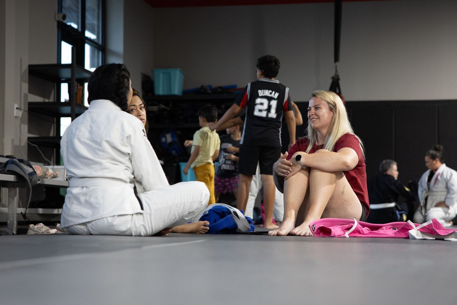 Two women sit on a gym floor chatting while athletes practice basketball in the background.