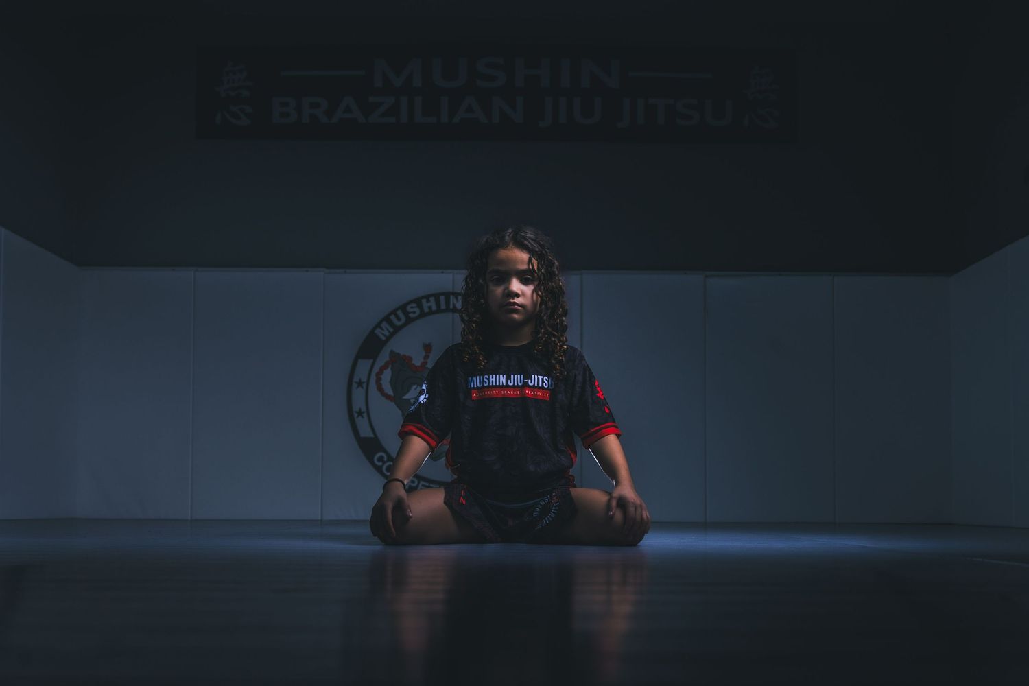 Young girl sits cross-legged on mat at Mushin Brazilian Jiu Jitsu gym in meditative pose.