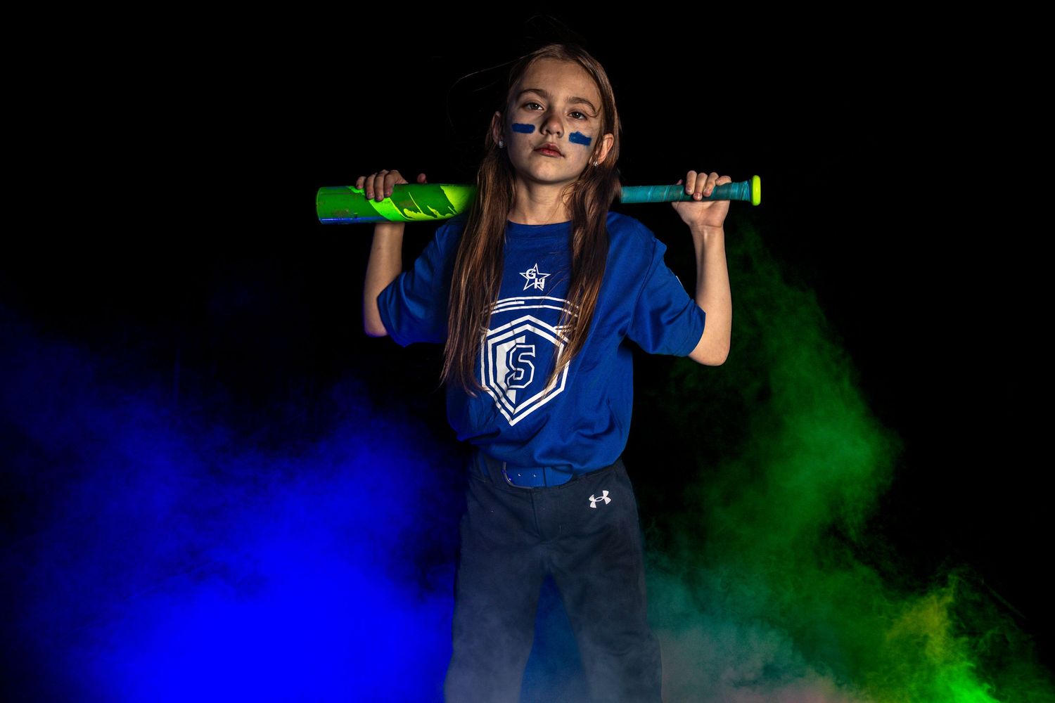 Young softball player poses with green bat on shoulders wearing blue jersey amid blue and green smoke on dark background.