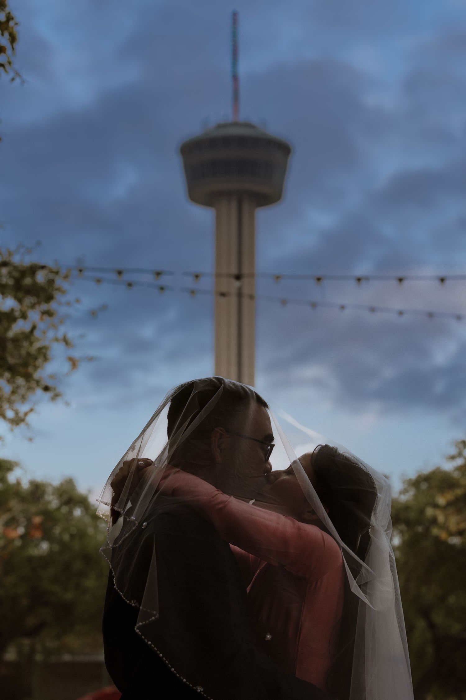 A romantic silhouette of a couple embracing at dusk with string lights and a tower in the background.