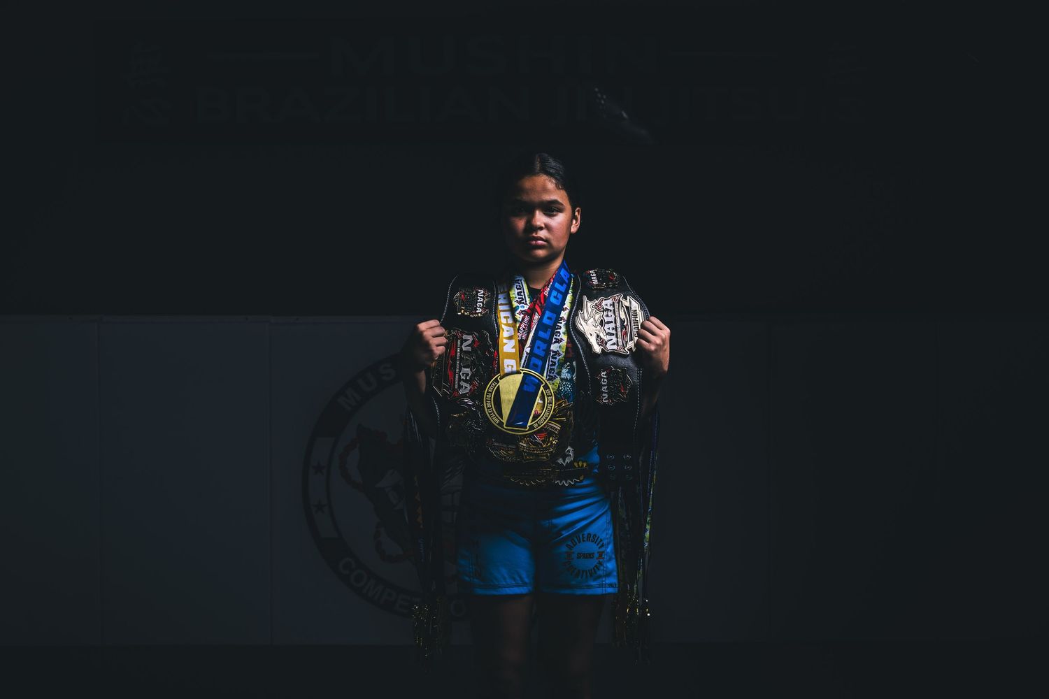 Young female martial artist proudly displays championship belt and medals in dramatic dark studio lighting.