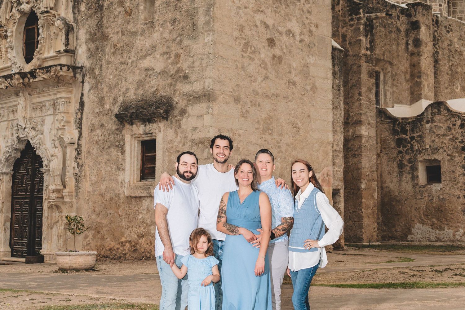 Family of six posing together outdoors in front of a historic stone mission building, dressed in coordinated blue and white outfits.