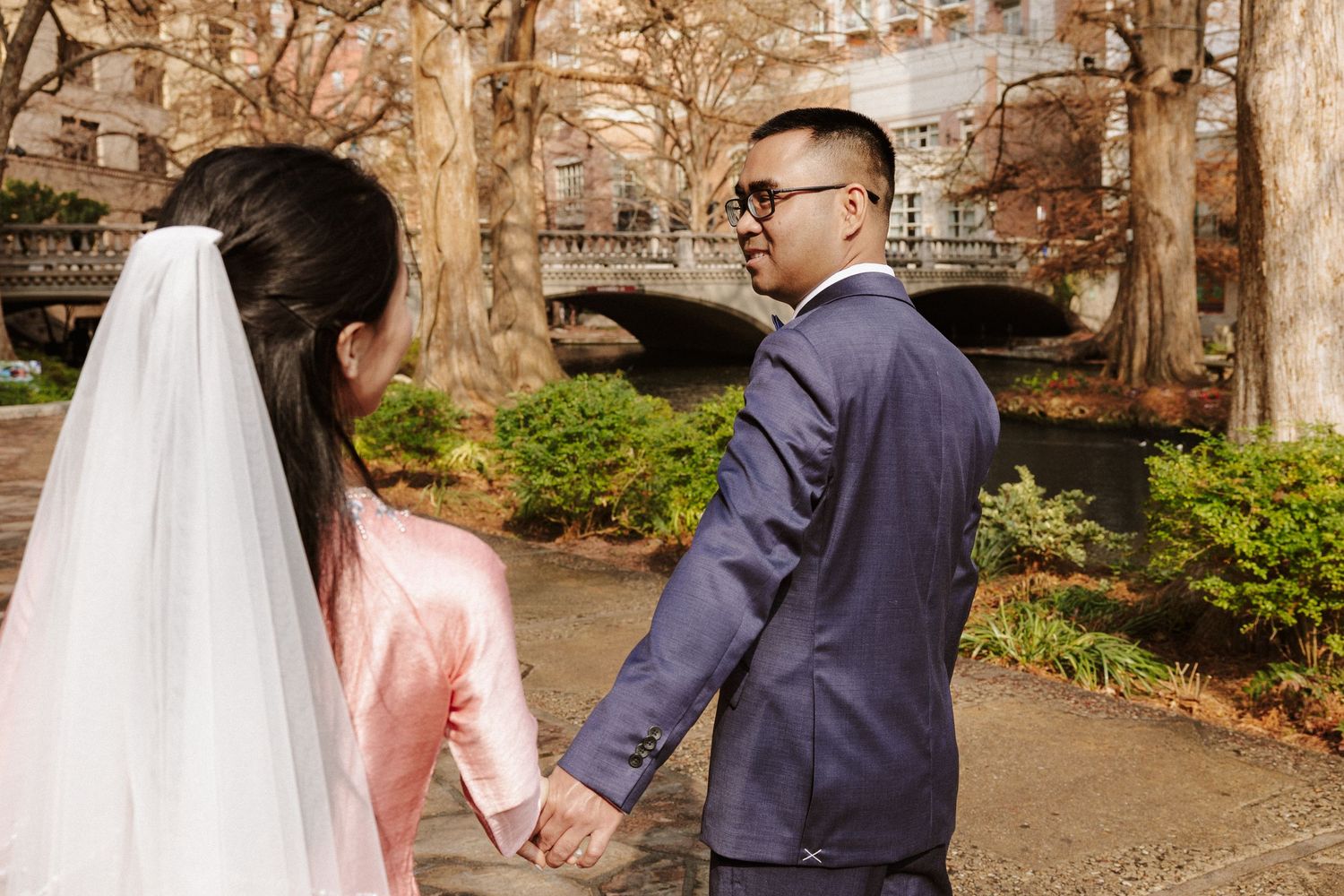 A couple in wedding attire walks through a scenic garden path with stone architecture in the background.