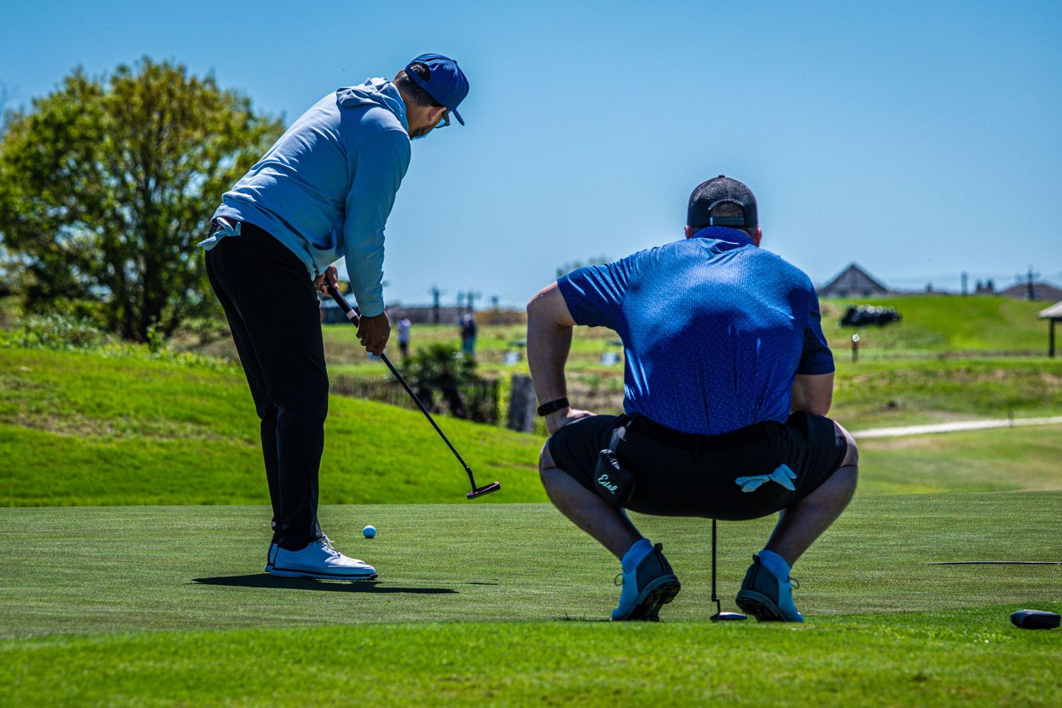 Two golfers on a putting green, one crouching to read the putt under a clear blue sky.