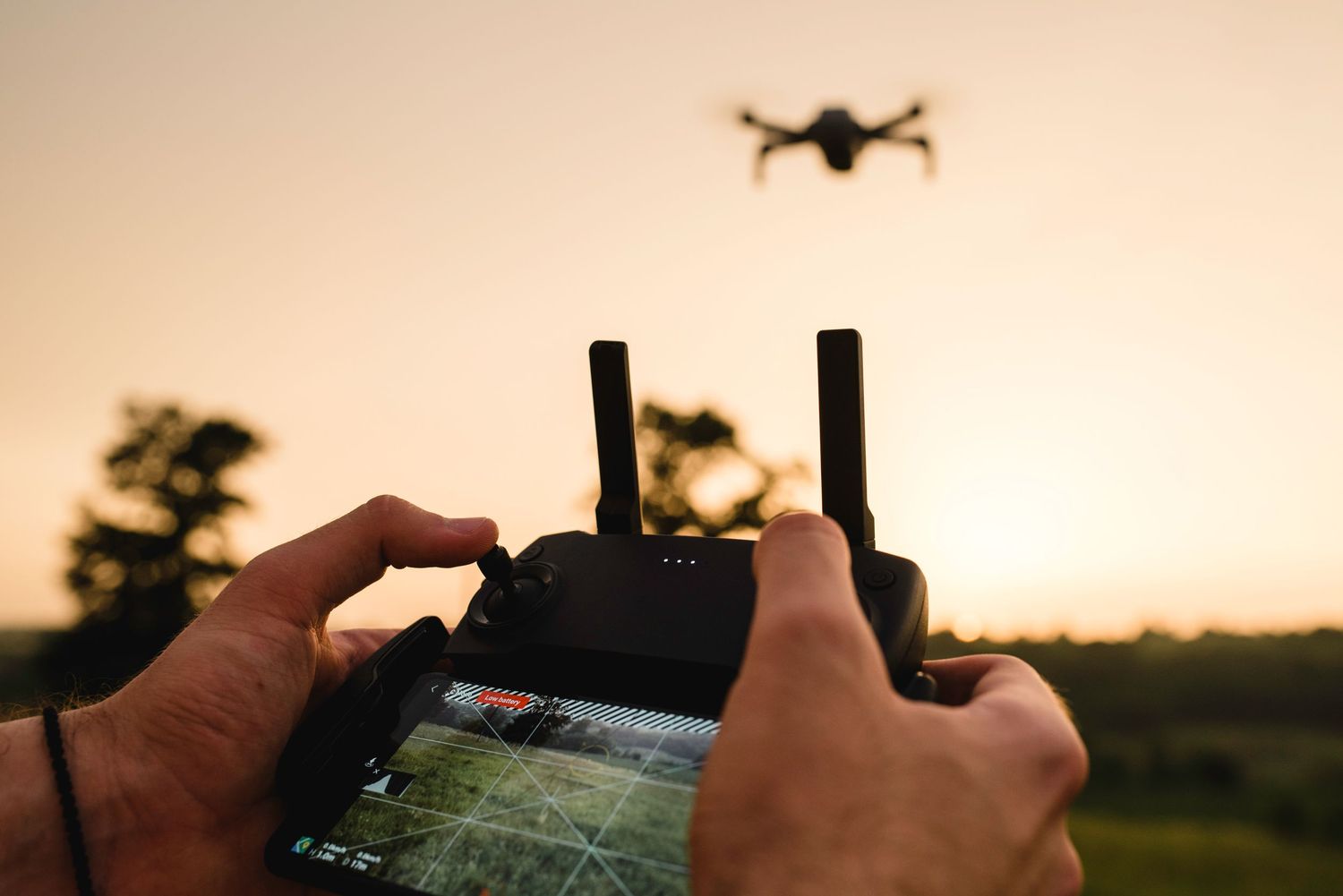 Person holding a drone remote controller with a quadcopter flying in the background at sunset.